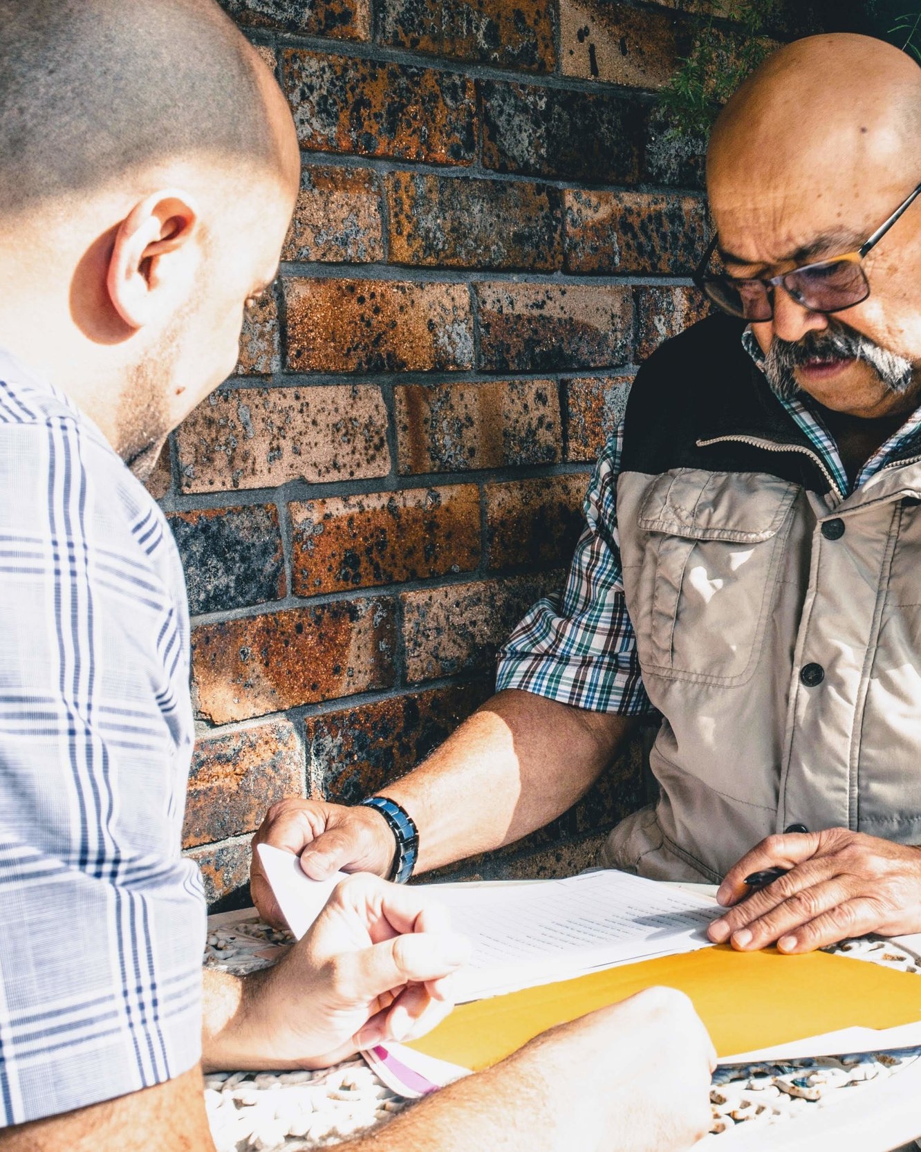 Two men collaborating on community-focused documents at an outdoor table, representing Telstra Health's co-design initiative. One man wears a plaid shirt and vest, while the other explains the papers. Brick wall and greenery in the background highlight a casual, approachable setting.nery in the background.