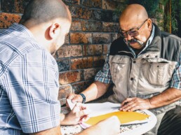 Two men collaborating on community-focused documents at an outdoor table, representing Telstra Health's co-design initiative. One man wears a plaid shirt and vest, while the other explains the papers. Brick wall and greenery in the background highlight a casual, approachable setting.nery in the background.