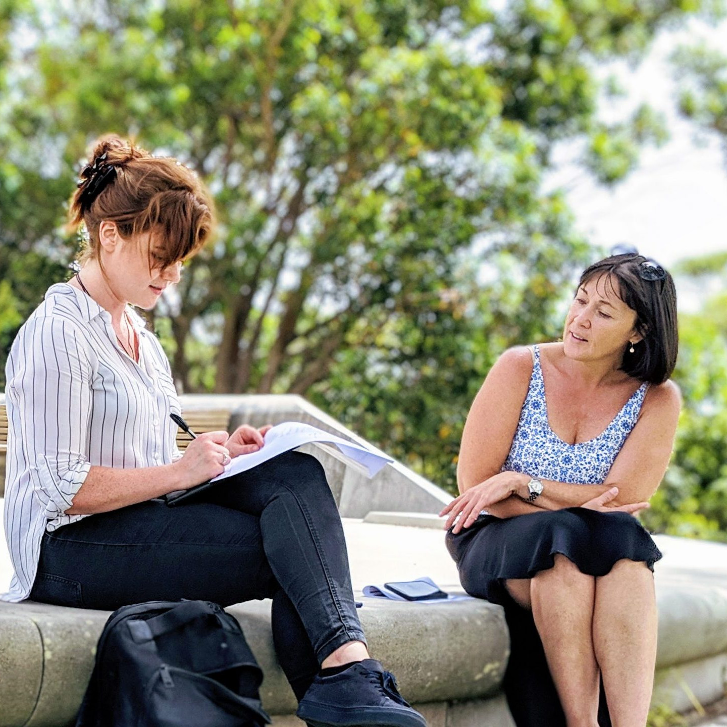 Two women sitting outdoors on a concrete bench in a green park, engaged in a conversation. One woman with red hair, wearing a striped shirt and black jeans, is writing notes in a notebook, while the other woman with dark hair, wearing a patterned sleeveless top and black skirt, listens attentively.