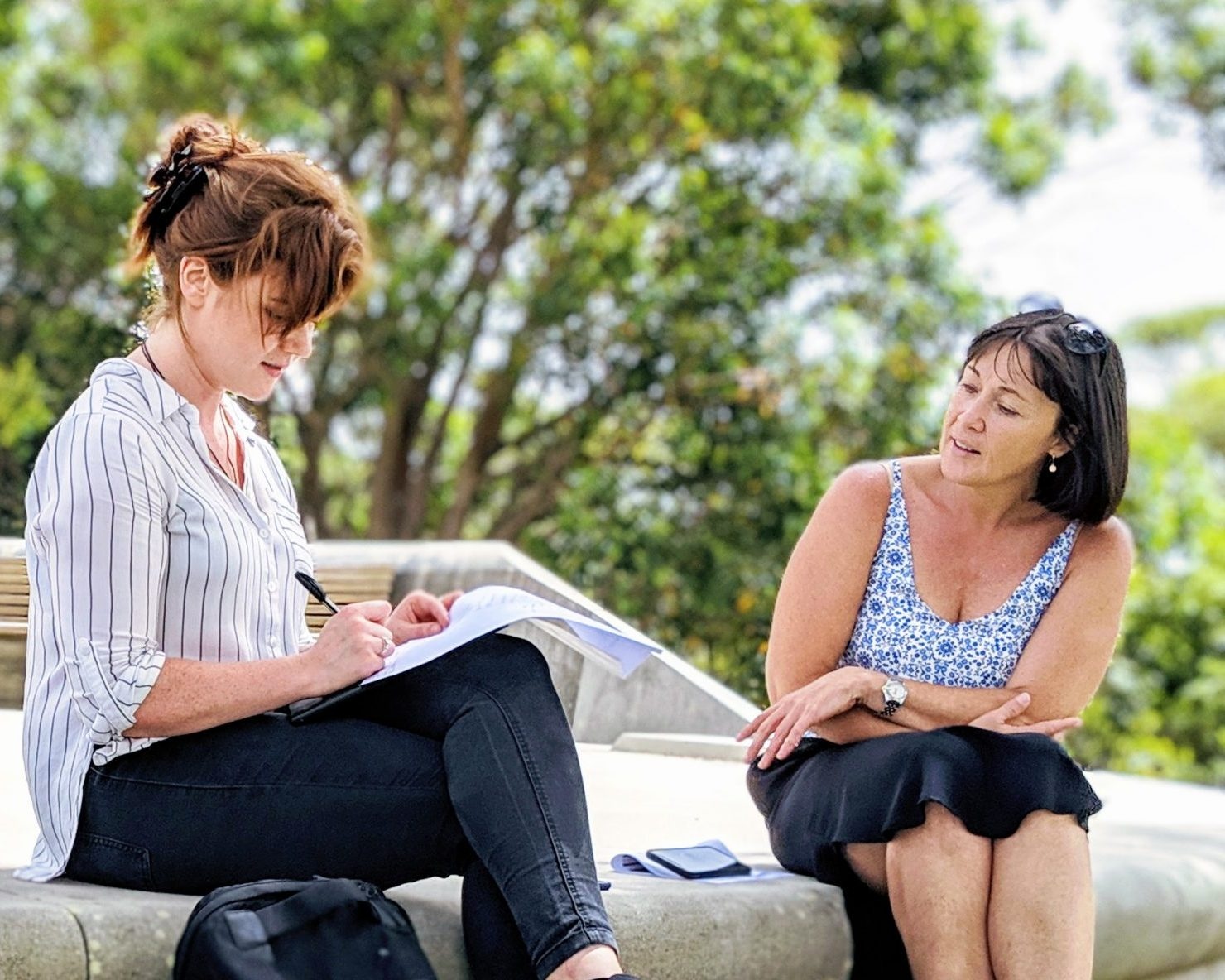 Two women sitting outdoors on a concrete bench in a green park, engaged in a conversation. One woman with red hair, wearing a striped shirt and black jeans, is writing notes in a notebook, while the other woman with dark hair, wearing a patterned sleeveless top and black skirt, listens attentively.