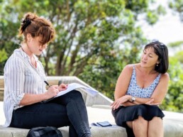 Two women sitting outdoors on a concrete bench in a green park, engaged in a conversation. One woman with red hair, wearing a striped shirt and black jeans, is writing notes in a notebook, while the other woman with dark hair, wearing a patterned sleeveless top and black skirt, listens attentively.