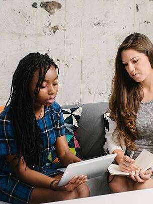 Two women seated on a sofa in a professional setting, one holding a tablet and the other taking notes in a notebook, with a coffee cup on a table.
