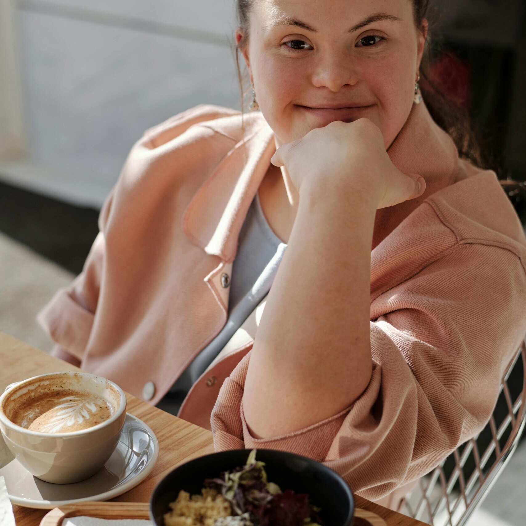 A young woman with Down syndrome smiling at the camera while sitting at a café table with a cup of coffee and a bowl of food.