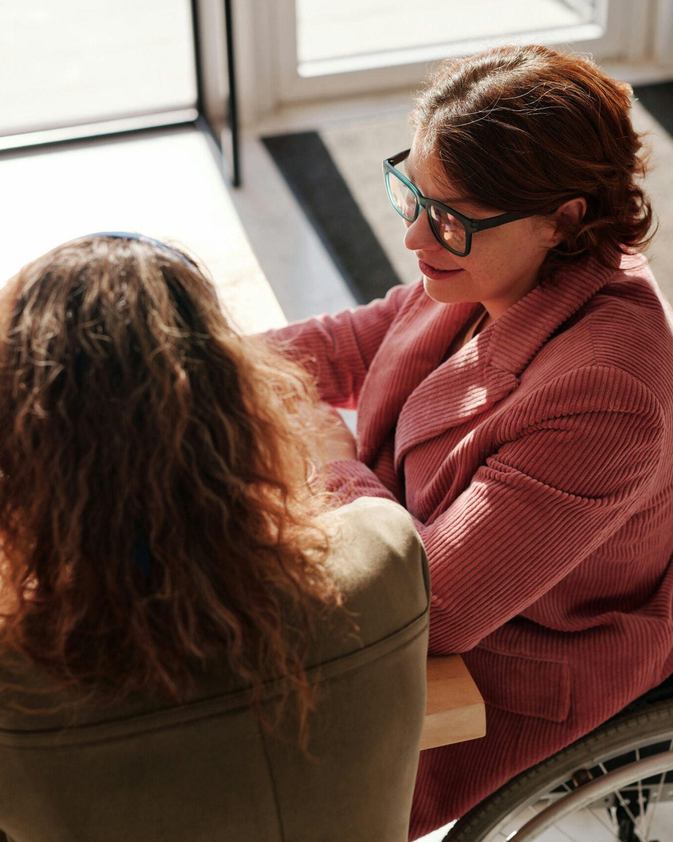 A woman in a wheelchair having a discussion with a colleague at a café table, with a laptop open and natural light streaming through the window.